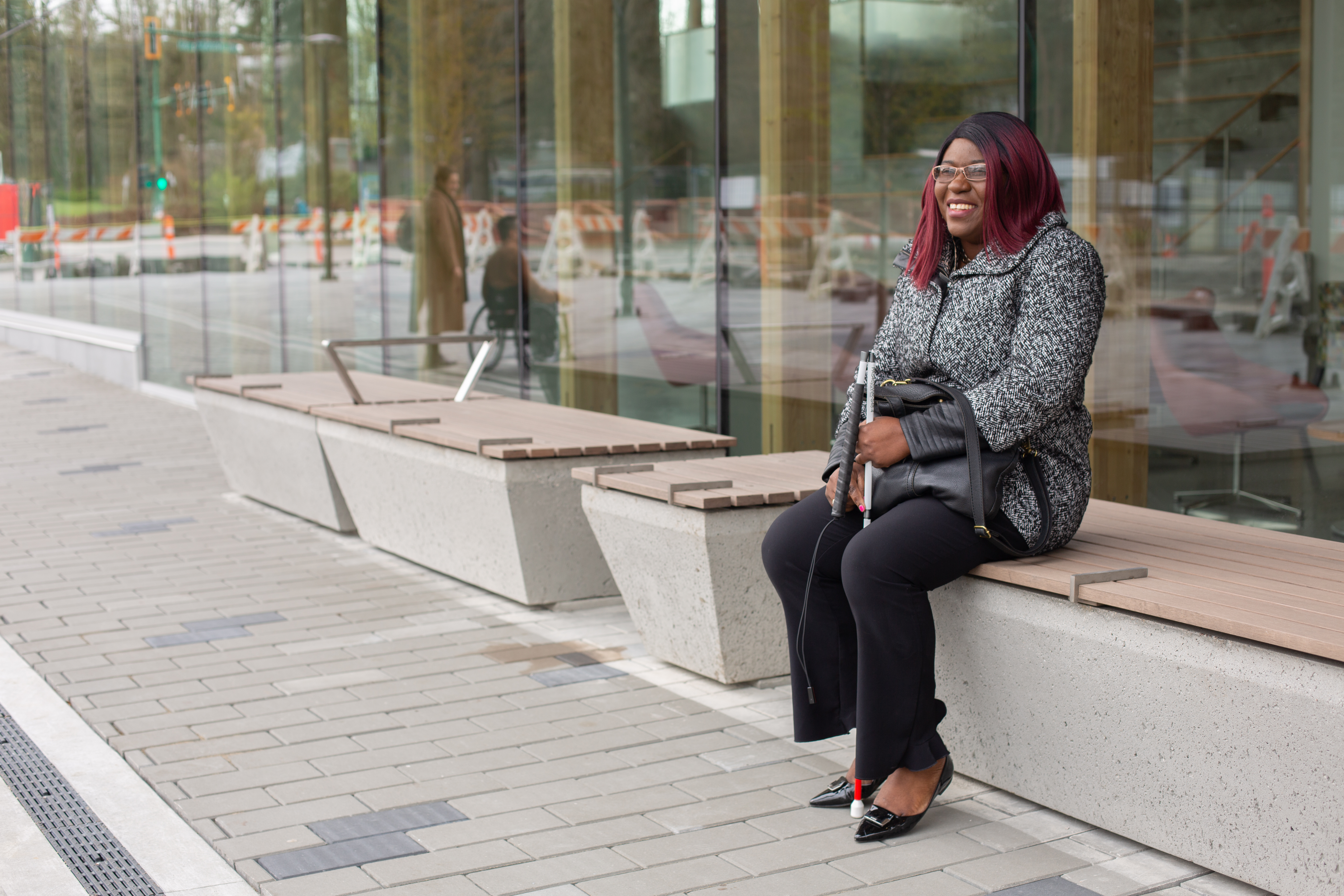 Laetitia Mfamobani smiling and sitting on a building bench outside wearing a black and white jacket and black pants. She has long pink and black hair and has a white cane.