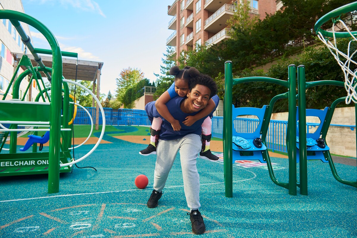 An adult and a young child in an accessible playground that is primarily blue and green. The child is on the adult's back, they are smiling.