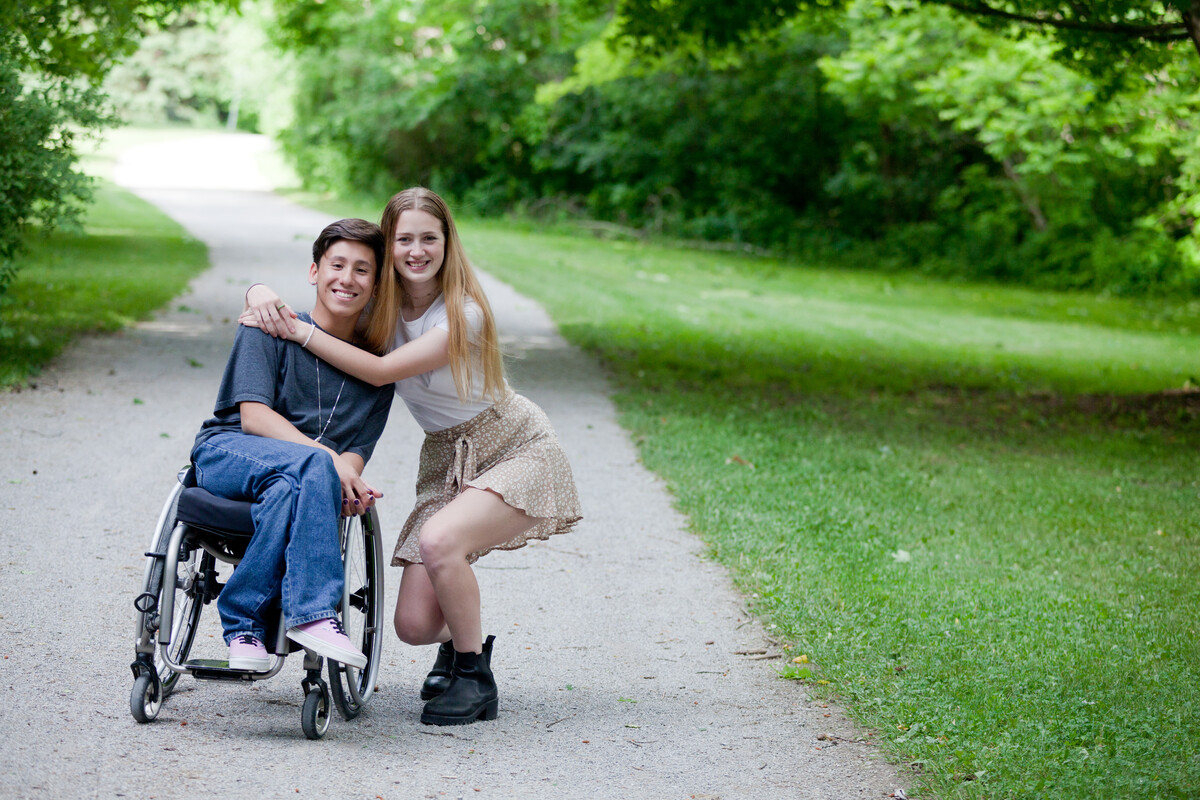 Two young people hugging on a path that is surrounded by trees. One person has short brown hair, is wearing jeans and is using a wheelchair. The other person has long hair, is wearing a skirt and is not using a wheelchair.