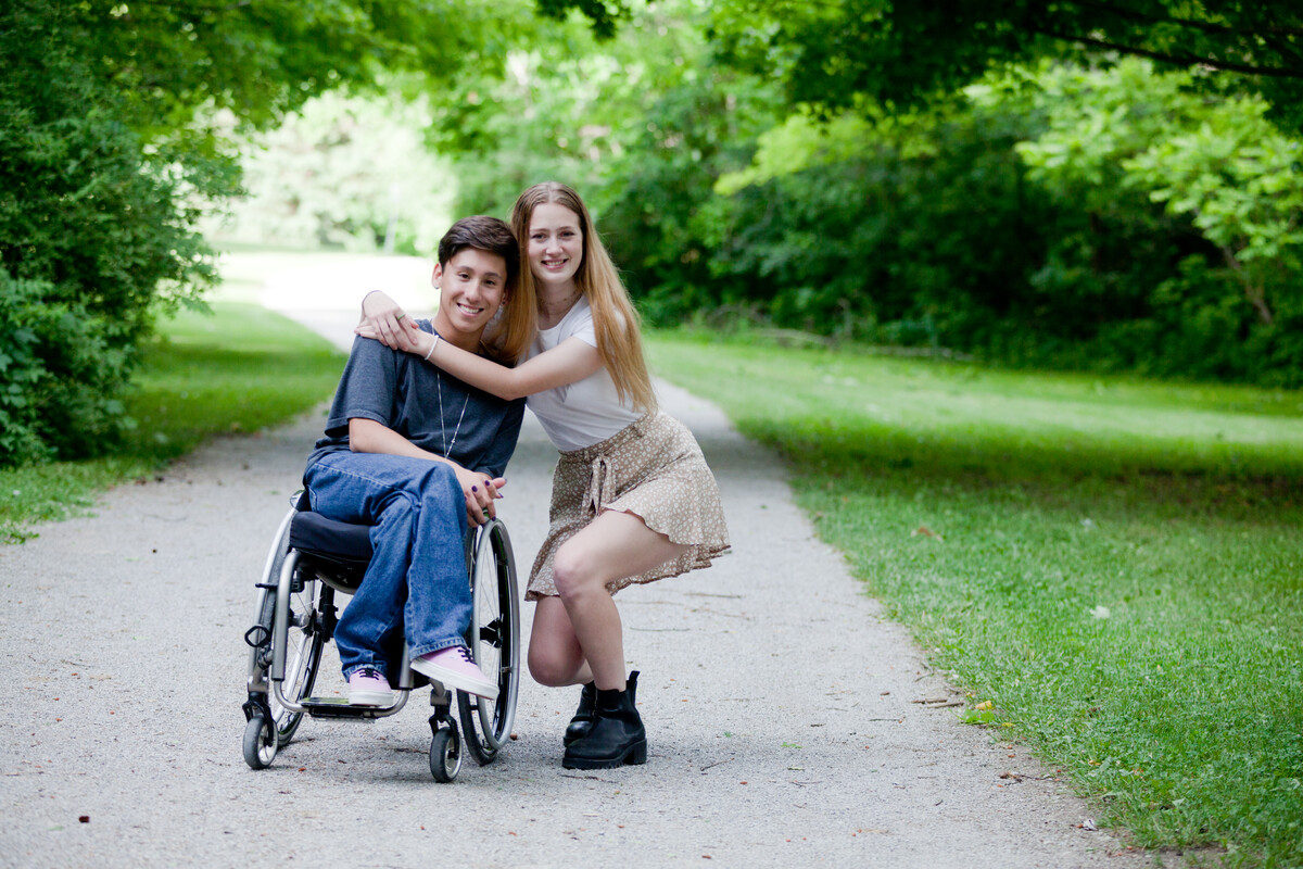 Two young people on a path outside, hugging and smiling at the camera. One person is using a wheelchair.