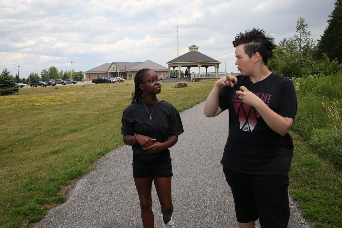 Two young people walking on a paved path in a park-like setting. One person with braided hair and glasses looks toward another person with short curly hair, who is communicating using sign language. A gazebo and community building are visible in the background.