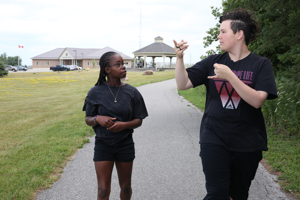 Two young people signing to each other. One person has long dark hair that is braided, the other person has short curly hair. They are both wearing black t-shirts.