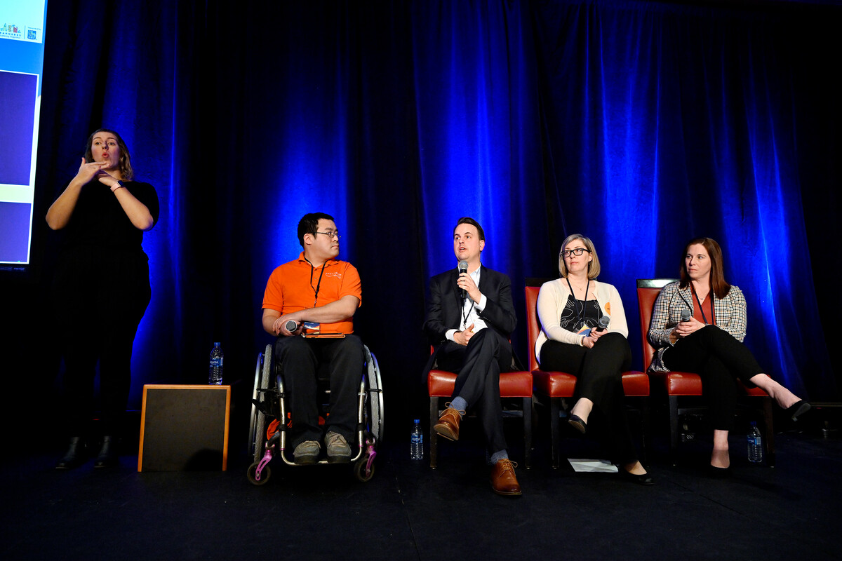 Panel discussion on a stage with blue curtains, featuring four speakers seated in chairs, including one person using a wheelchair, and a sign language interpreter standing to the side.