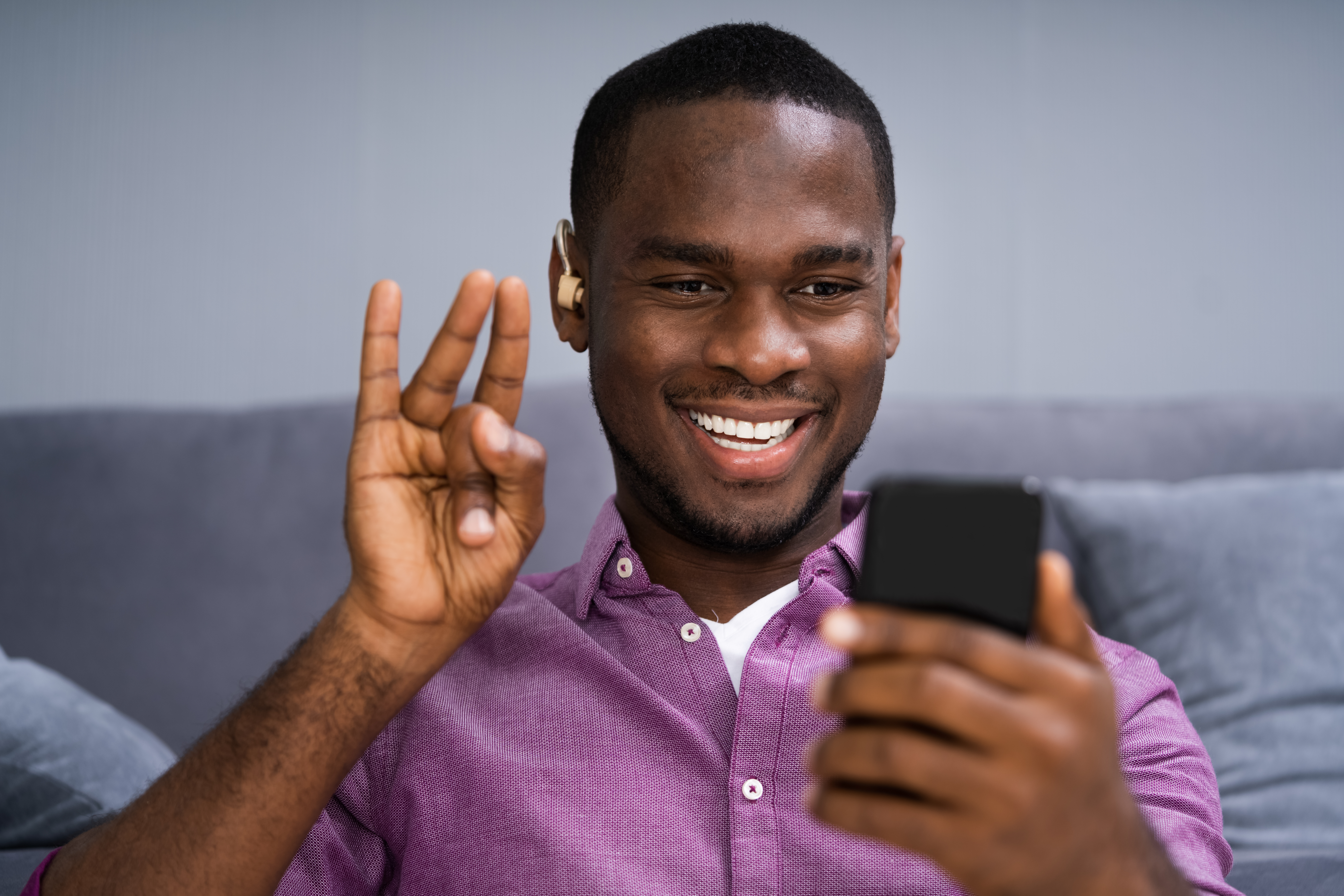 A man wearing a hearing aid smiles at his phone in his left hand while signing with his right hand