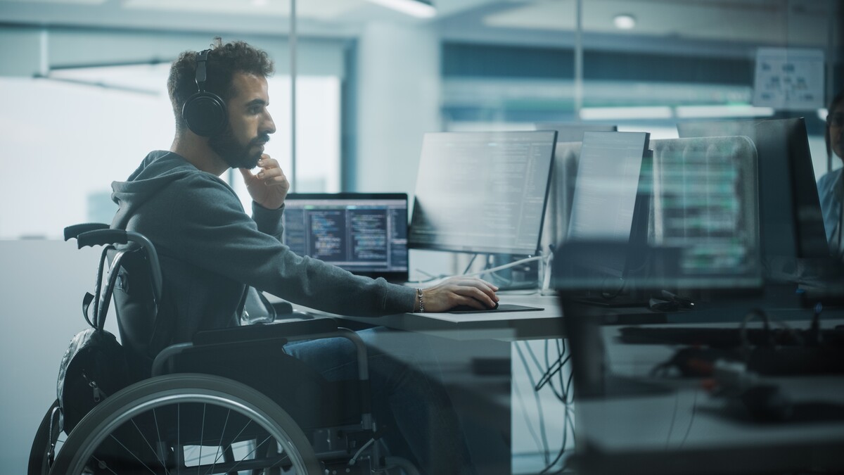 Person with short dark hair wearing headphones who is using a wheelchair at a computer desk with three computer monitors. 