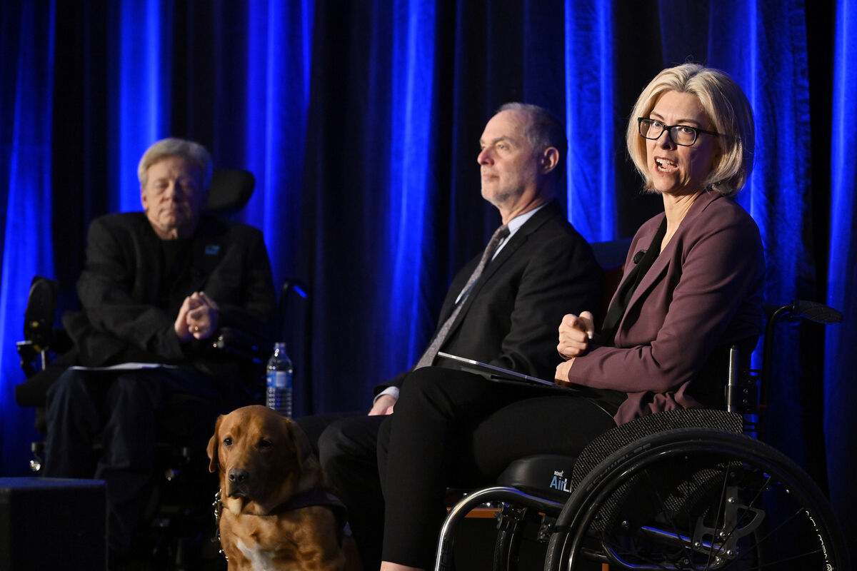 Cadieux using a wheelchair in a nanel discussion on a stage, with other two speakers and a guide dog