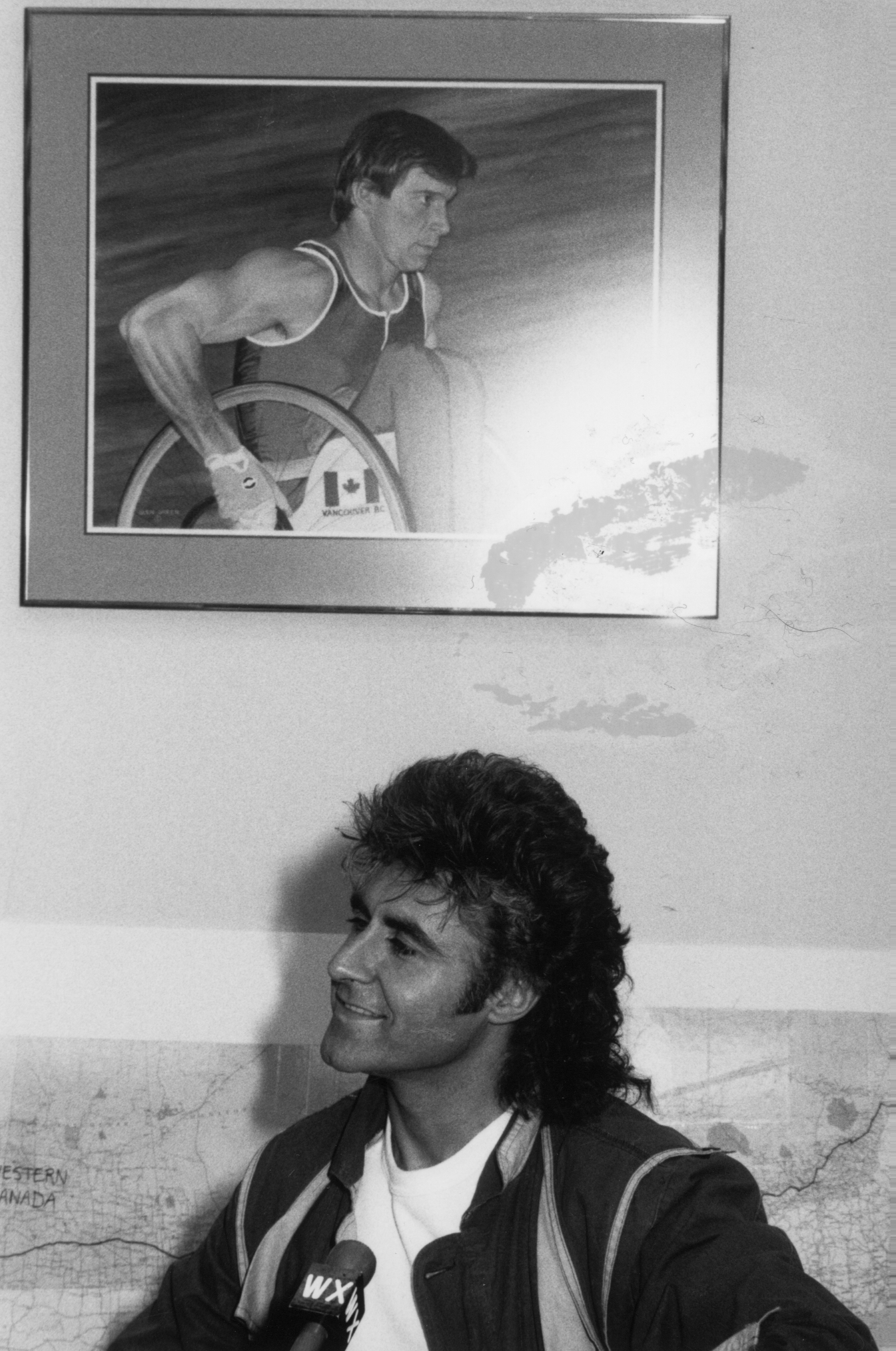 A man sitting on a couch and holding a microphone. Behind the person, there is a framed picture on the wall of an athlete pushing the wheel of a wheelchair. The athlete's (Rick) uniform features a maple leaf