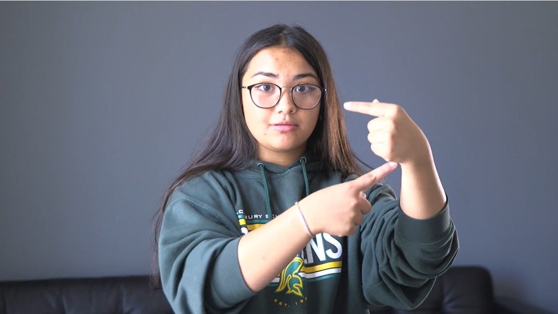 Young woman wearing glasses and a hoodie, using both hands to form a rectangular frame gesture against a plain wall background.
