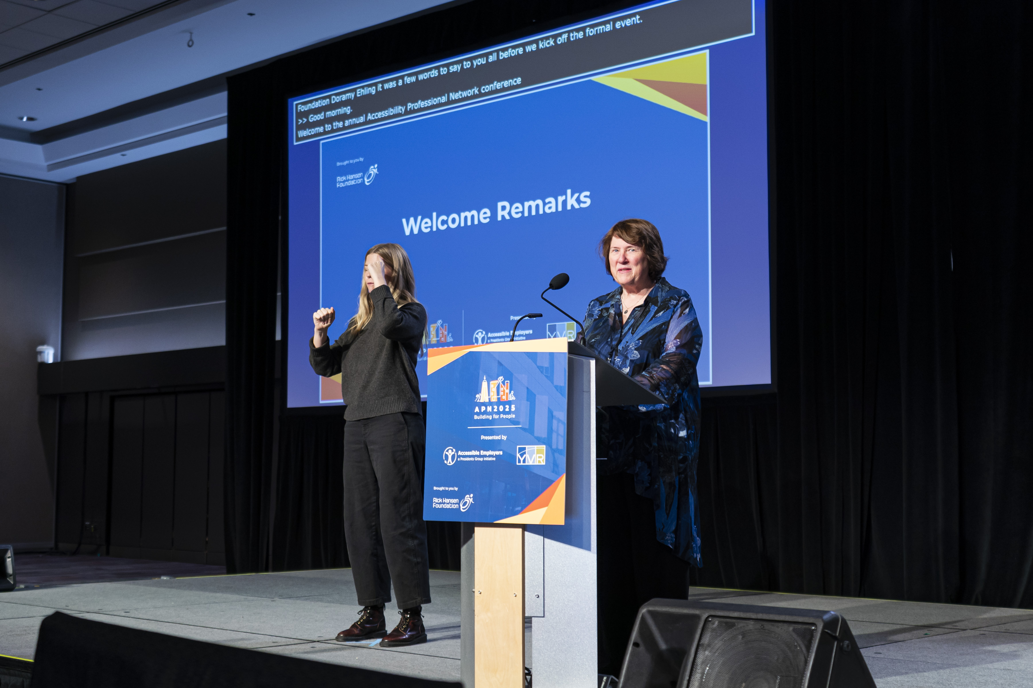 Two individuals are on stage: one, a woman, stands at a podium with "Welcome Remarks" displayed on a large blue screen behind her, while the other person is using sign language to interpret.