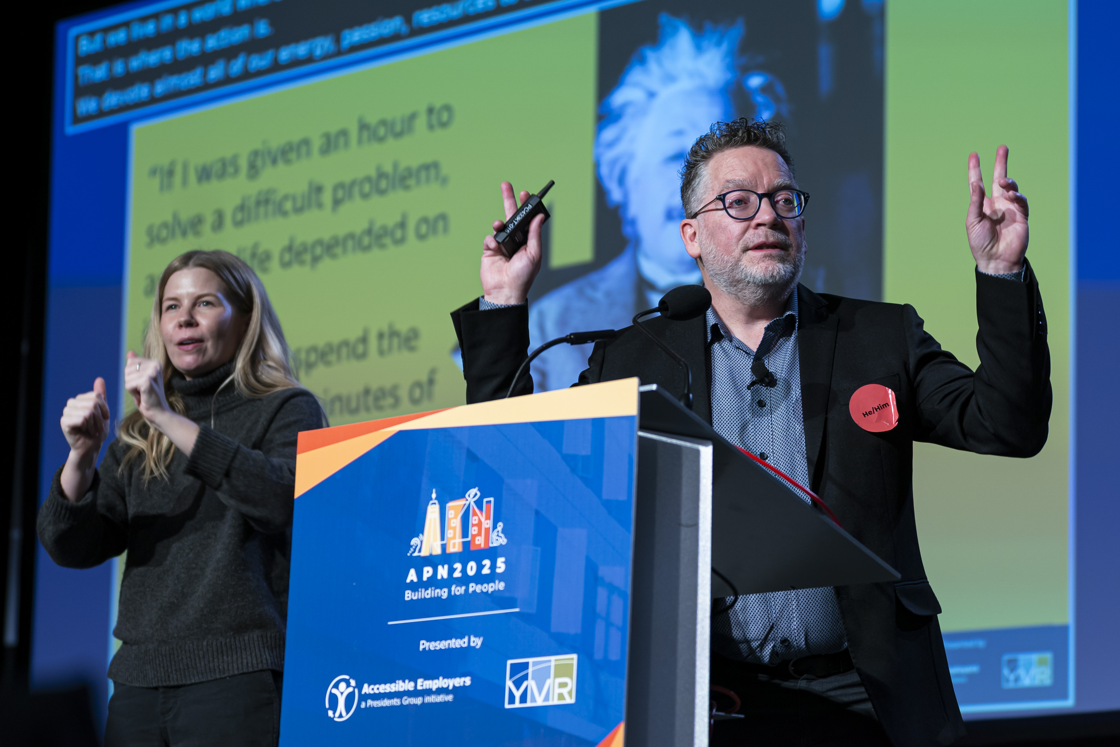 On stage, two individuals:a man delivers his presentation while a woman interprets using sign language. Behind them, a large screen with a yellow background, displaying black font and live captions