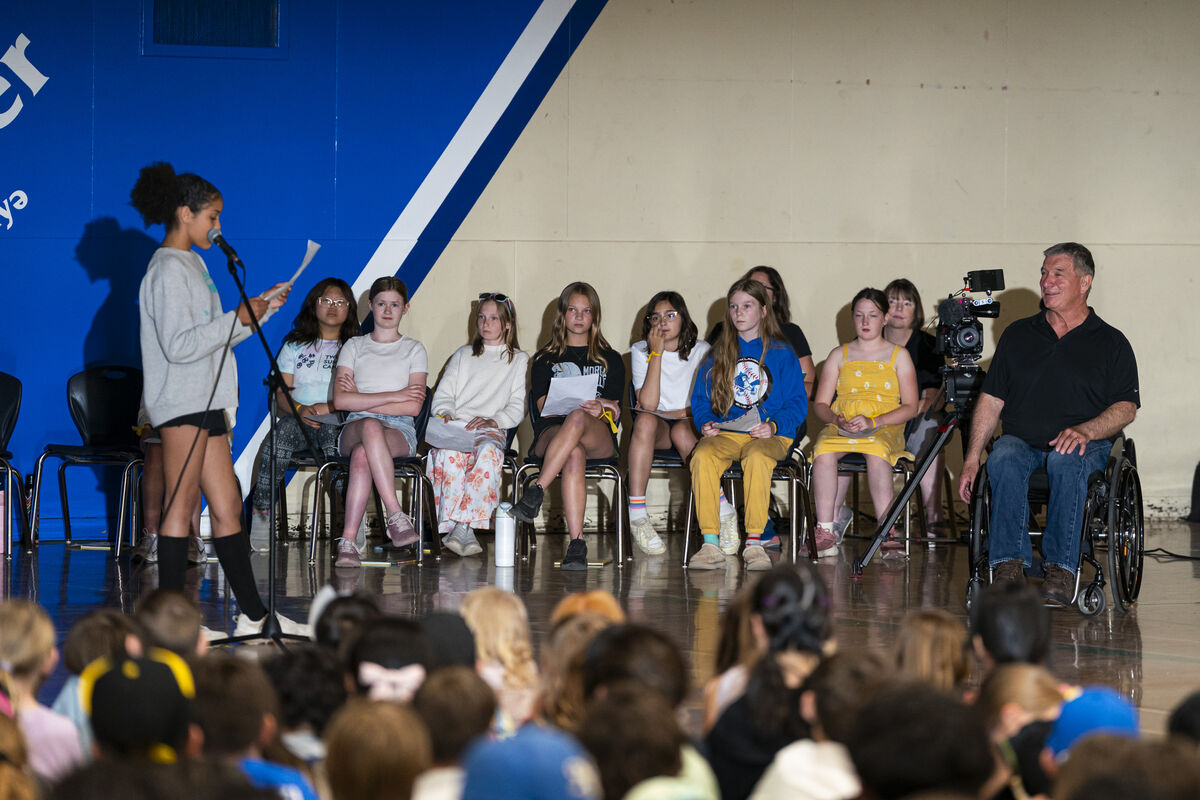 Student standing at a microphone reading from a sheet of paper. Several students are seated behind them on stage, while a person seated in a wheelchair and a video camera are positioned nearby.