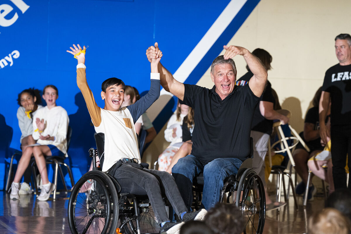 Young boy and a man seated in wheelchairs raise their joined hands during a school assembly, with students seated on stage in the background.
