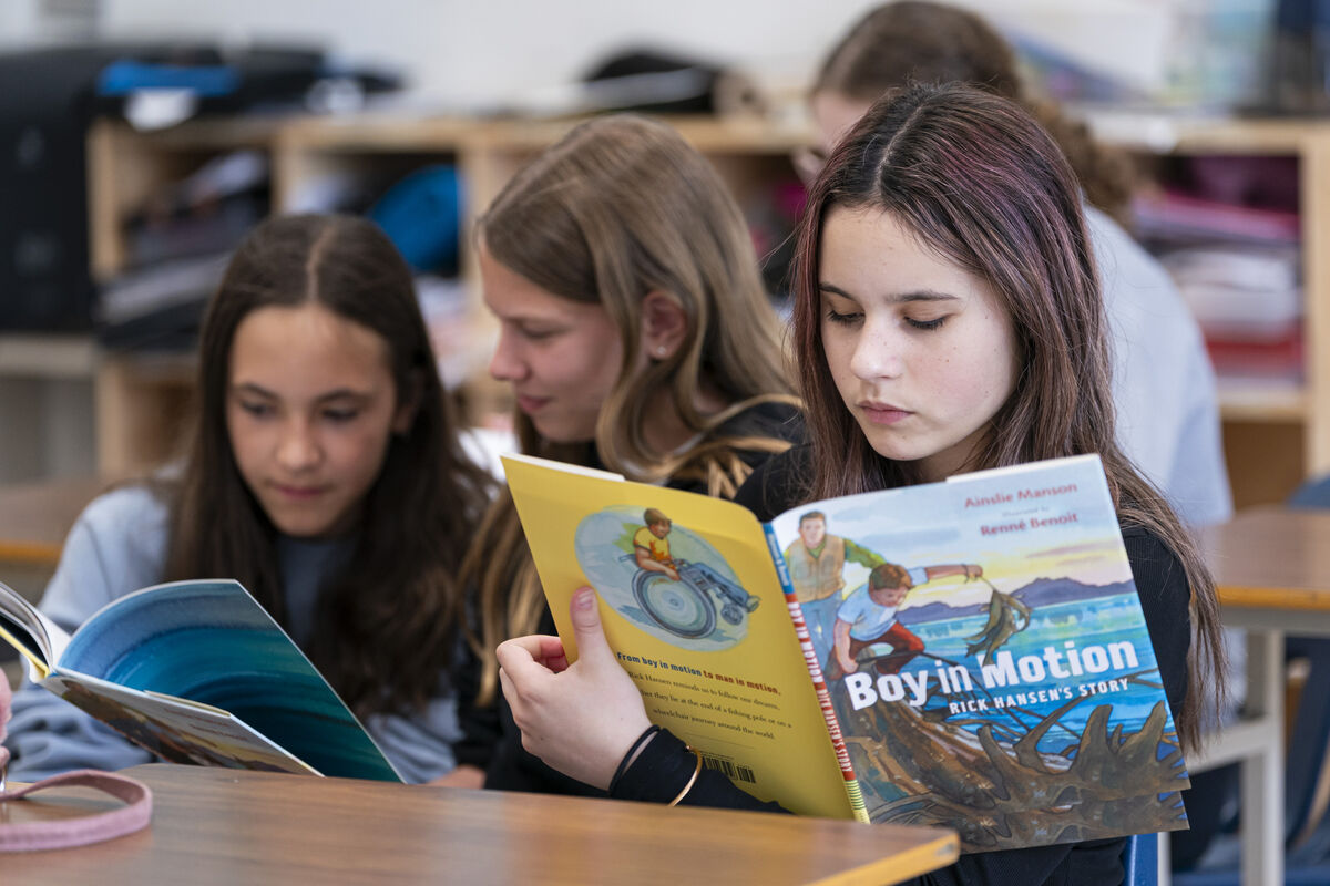 Students seated at a classroom table reading books. One student is reading Boy in Motion: Rick Hansen's Story while others read nearby.