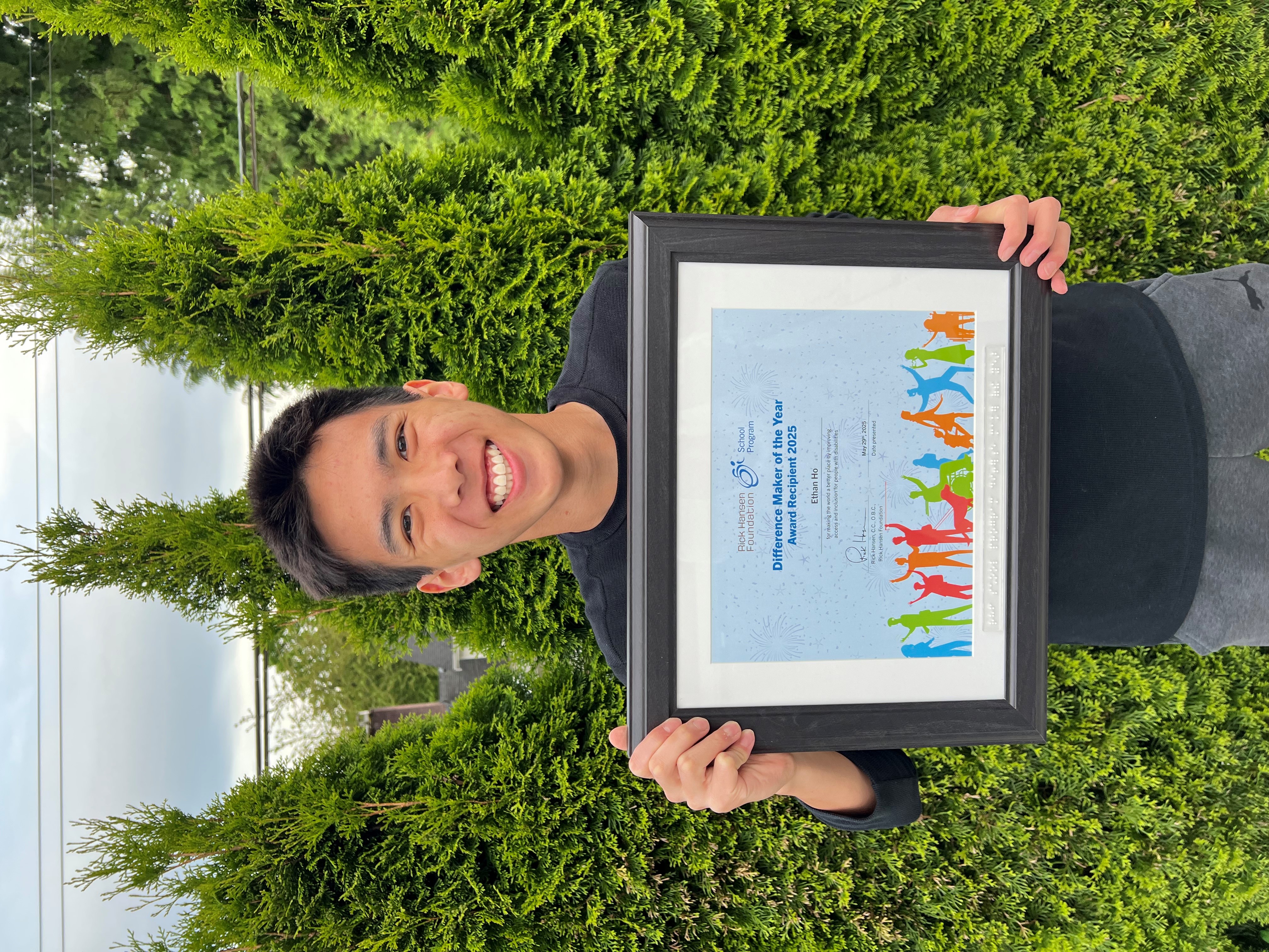 A young Asian boy standing outdoors in front of tall green shrubs, holding a framed certificate with both hands. The certificate reads “Difference Maker Award 2025” and the Rick Hansen Foundation logo.