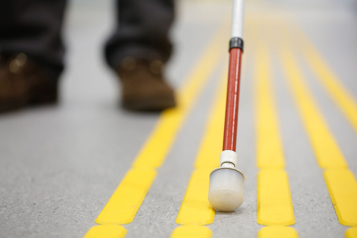 Close-up of a white cane with a red section moving along bright yellow tactile guiding strips on the ground.