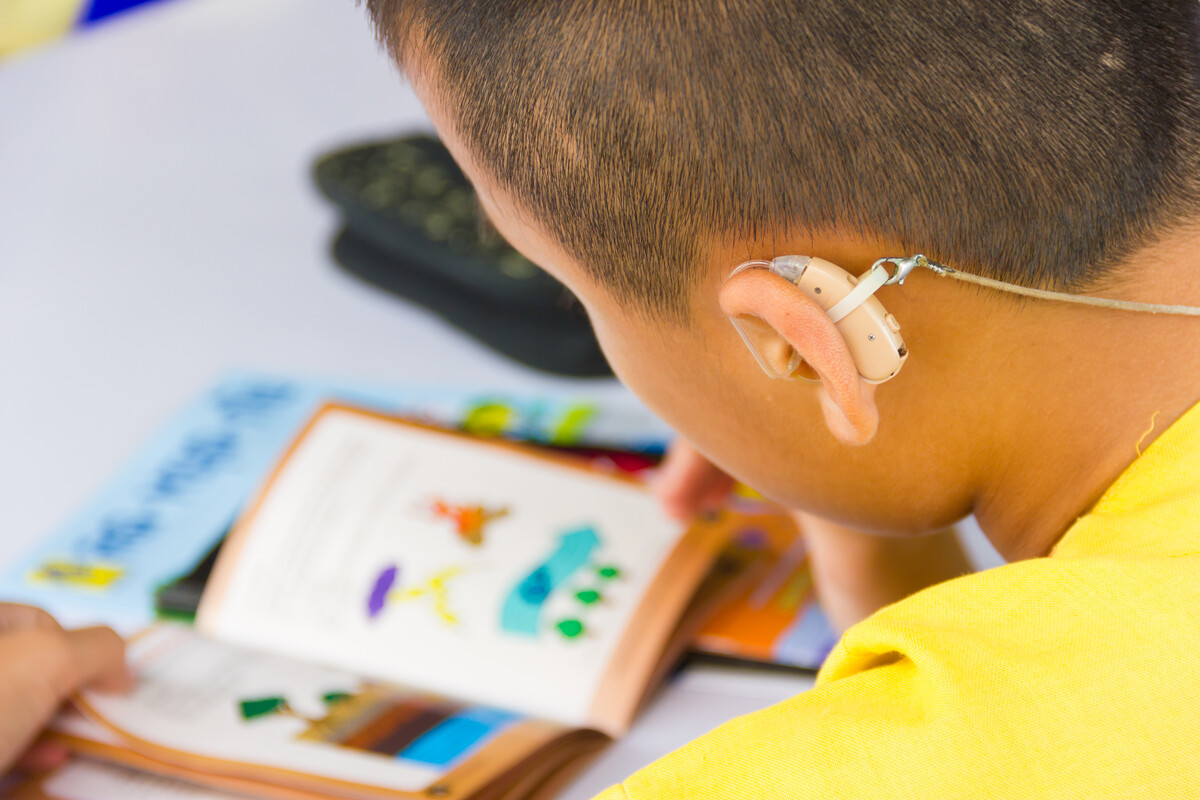 Close-up of a child wearing a hearing aid reading an illustrated book at a table.
