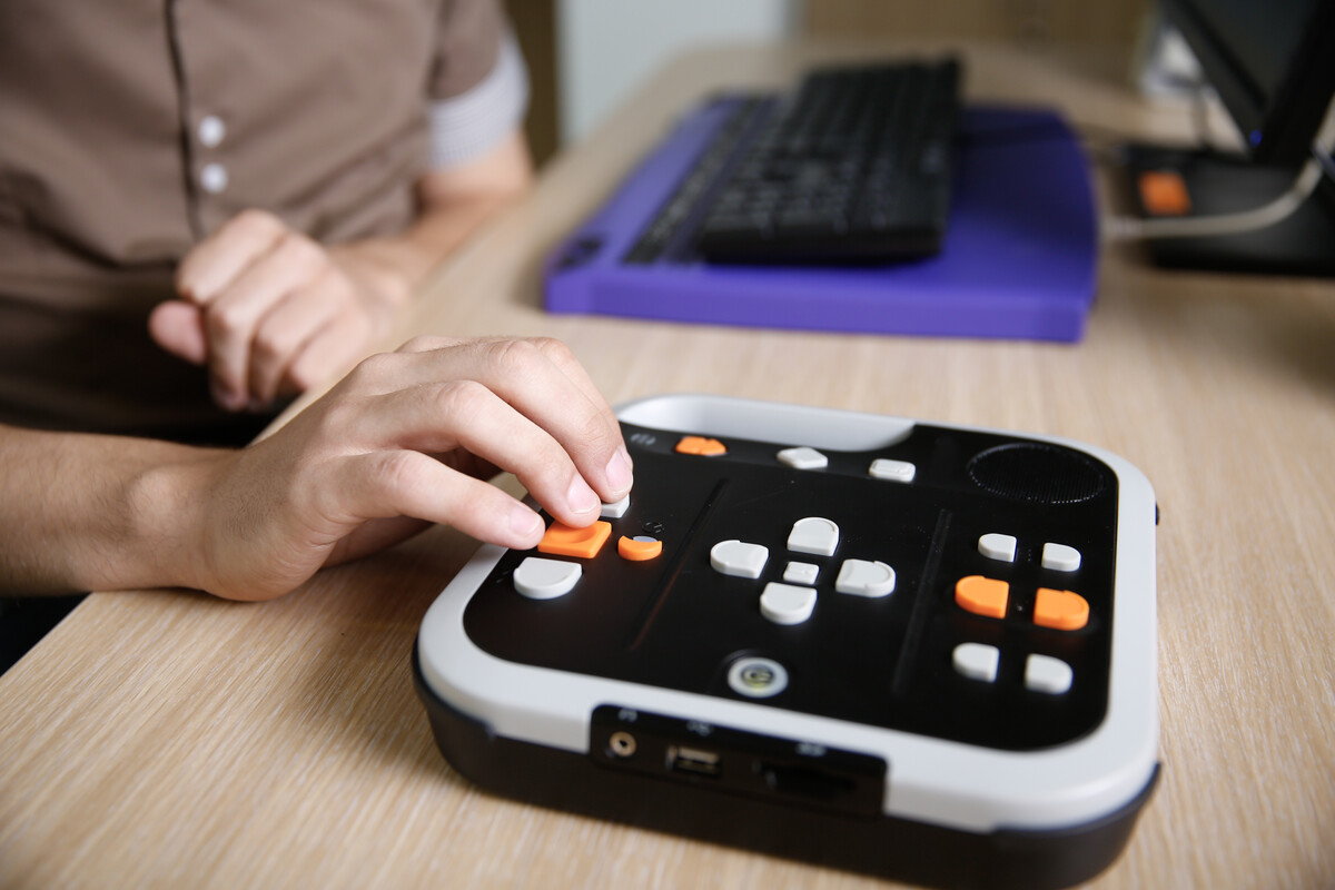 Person using an assistive communication device with large buttons on a table, with a keyboard and computer in the background.