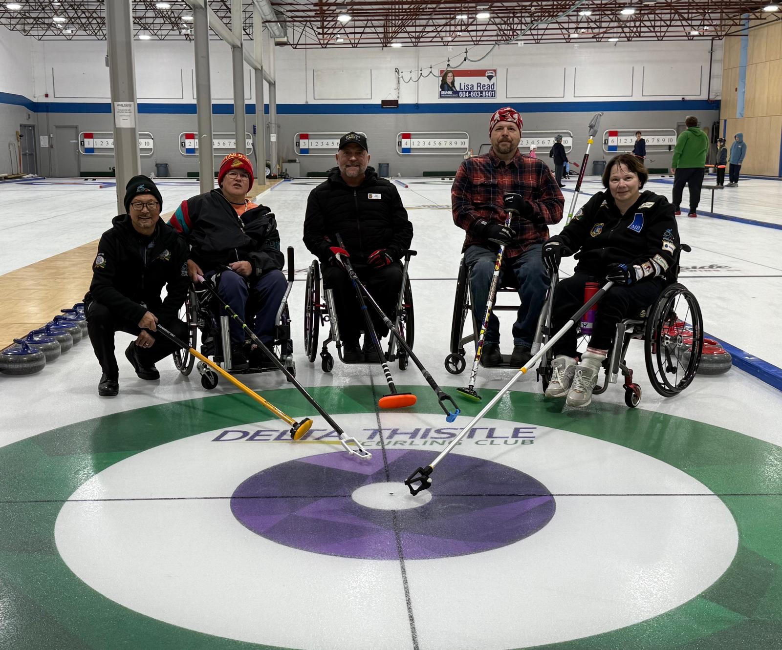 Five wheelchair curlers posing together on the ice at the center of a curling house. Each person holds a delivery stick pointing toward the target circle. Curling stones and equipment are visible along the side.