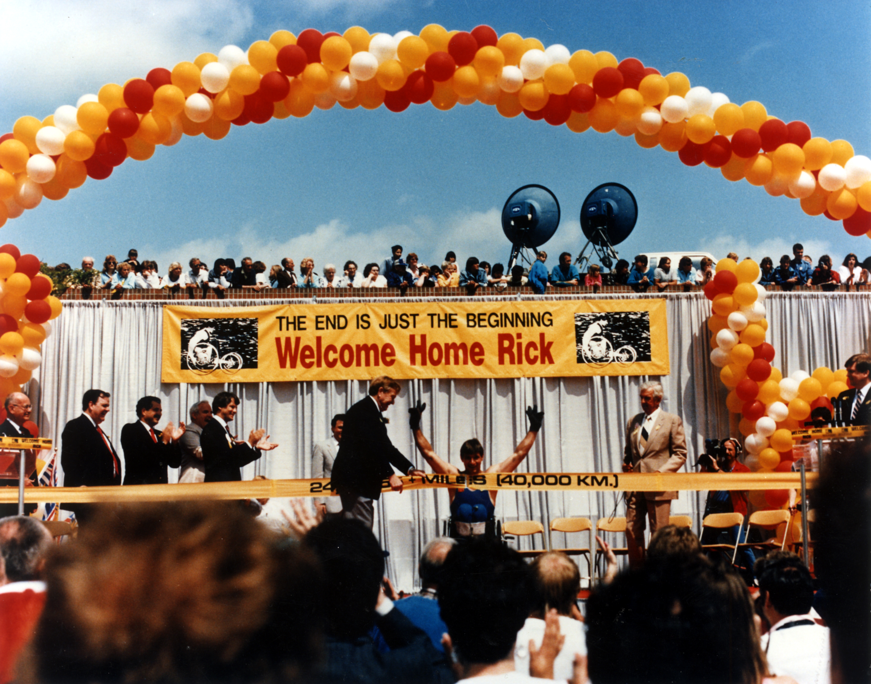 A celebratory event shows a large crowd gathered in front of a stage decorated balloons. On the stage, several people as a man in a wheelchair, wearing athletic gear and raising his arms crosses under a yellow ribbon that reads '40,000 KM.' Above the stage is a large banner stating 'THE END IS JUST THE BEGINNING Welcome Home Rick'