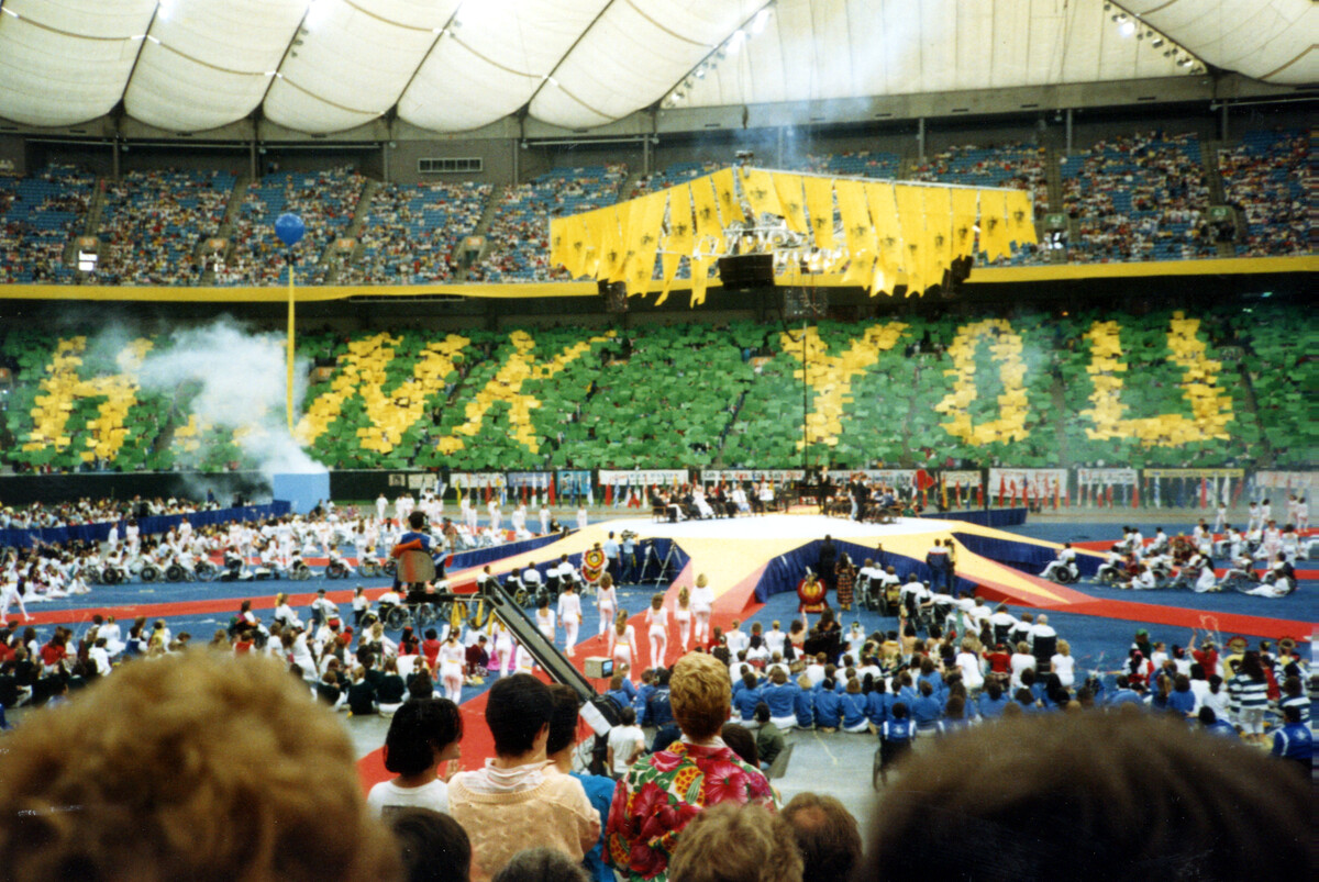 An arena filled with cheers fans for Rick Hansen following the end of the World Tour. In the stands, fans have created the words "Thank you" using yellow posters.