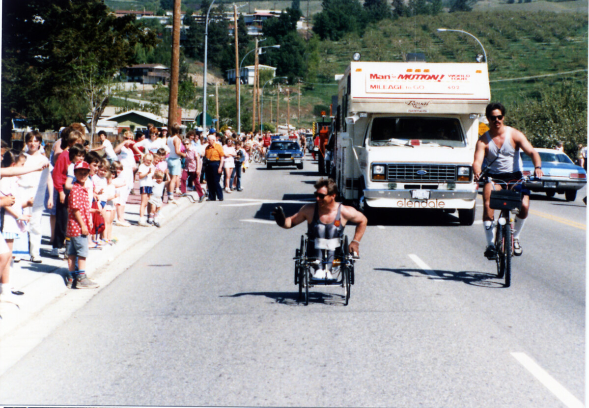 The Man In Motion World Tour. Rick is wheeling down the street with his motorhome close behind. He is surrounded by supporters.