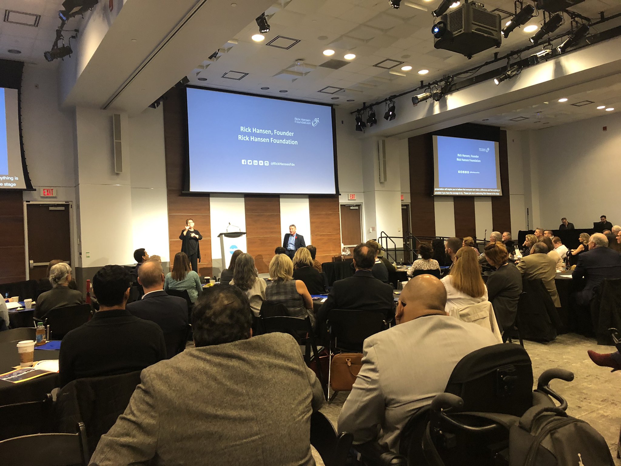 People in a conference room looking at Rick Hansen who is presenting at the front of the room. There is an ASL interpreter and a projector screen that reads "Rick Hansen, Founder Rick Hansen Foundation"