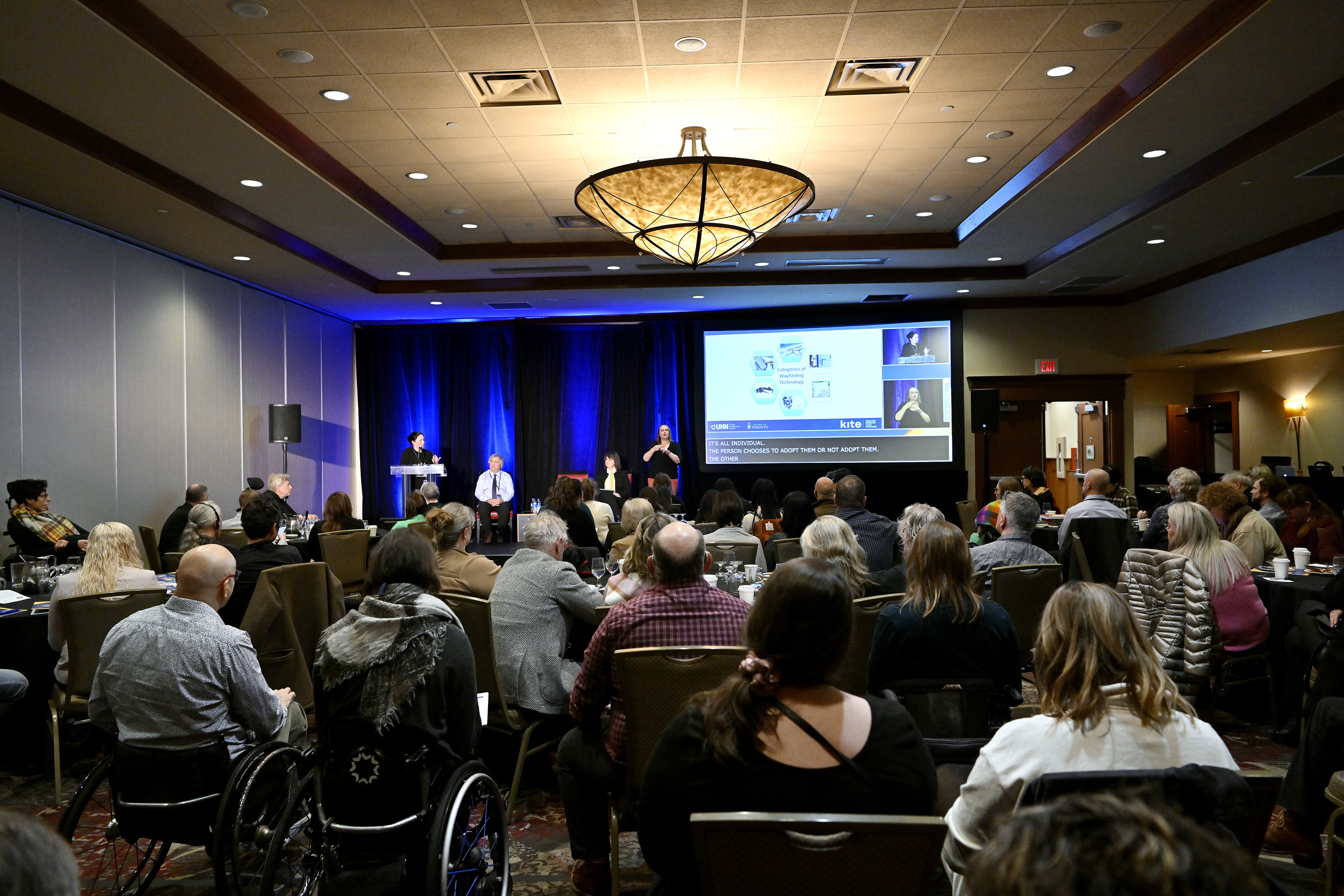 A crowd of people at a conference watching people on stage. There is a Power Point presentation and an ASL interpreter present.