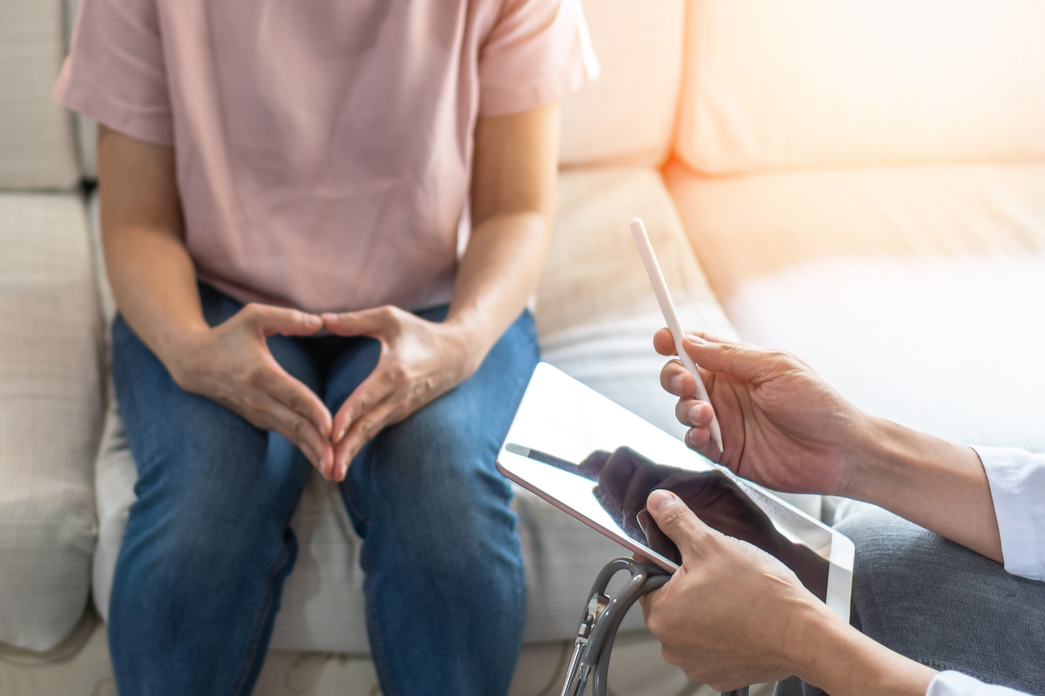A seated woman with hands clasped together. In front of her, another individual holds a digital tablet and a pen.