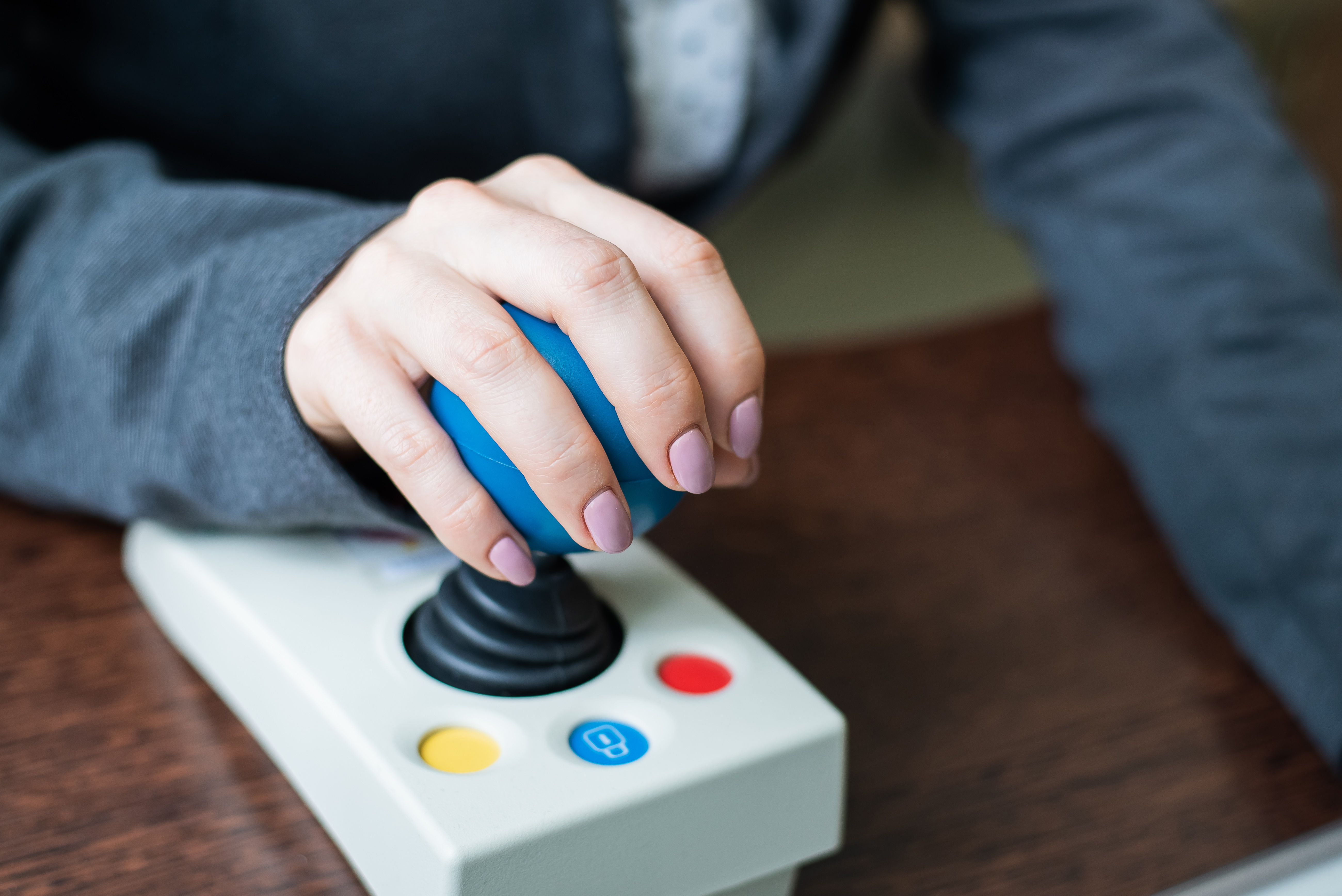 Close-up of a hand gripping a joystick controller with coloured buttons on an assistive device placed on a desk.