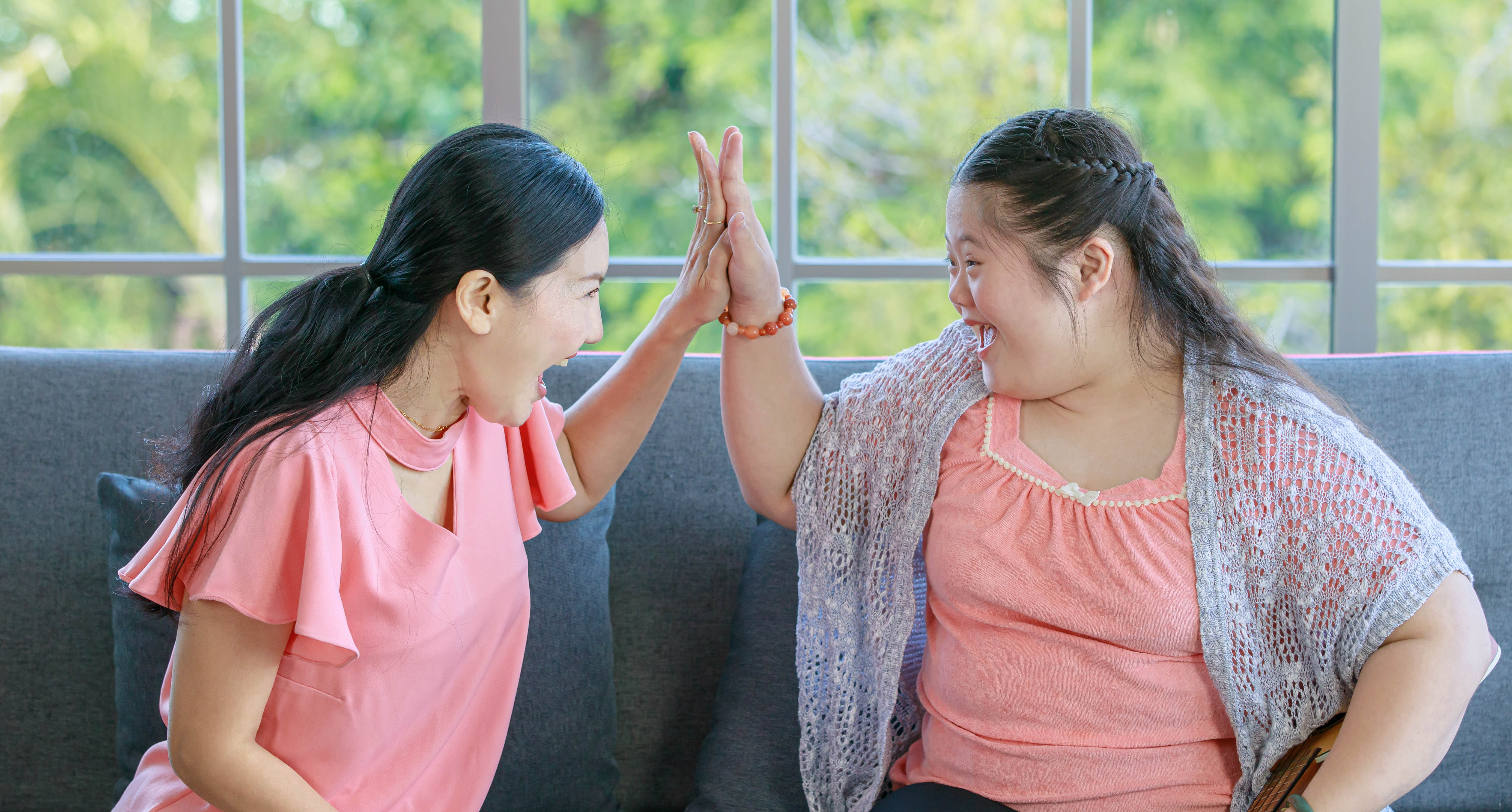 Two woman sit on a couch indoors and raise their hands to meet in a high‑five, with a large window and greenery visible behind them.