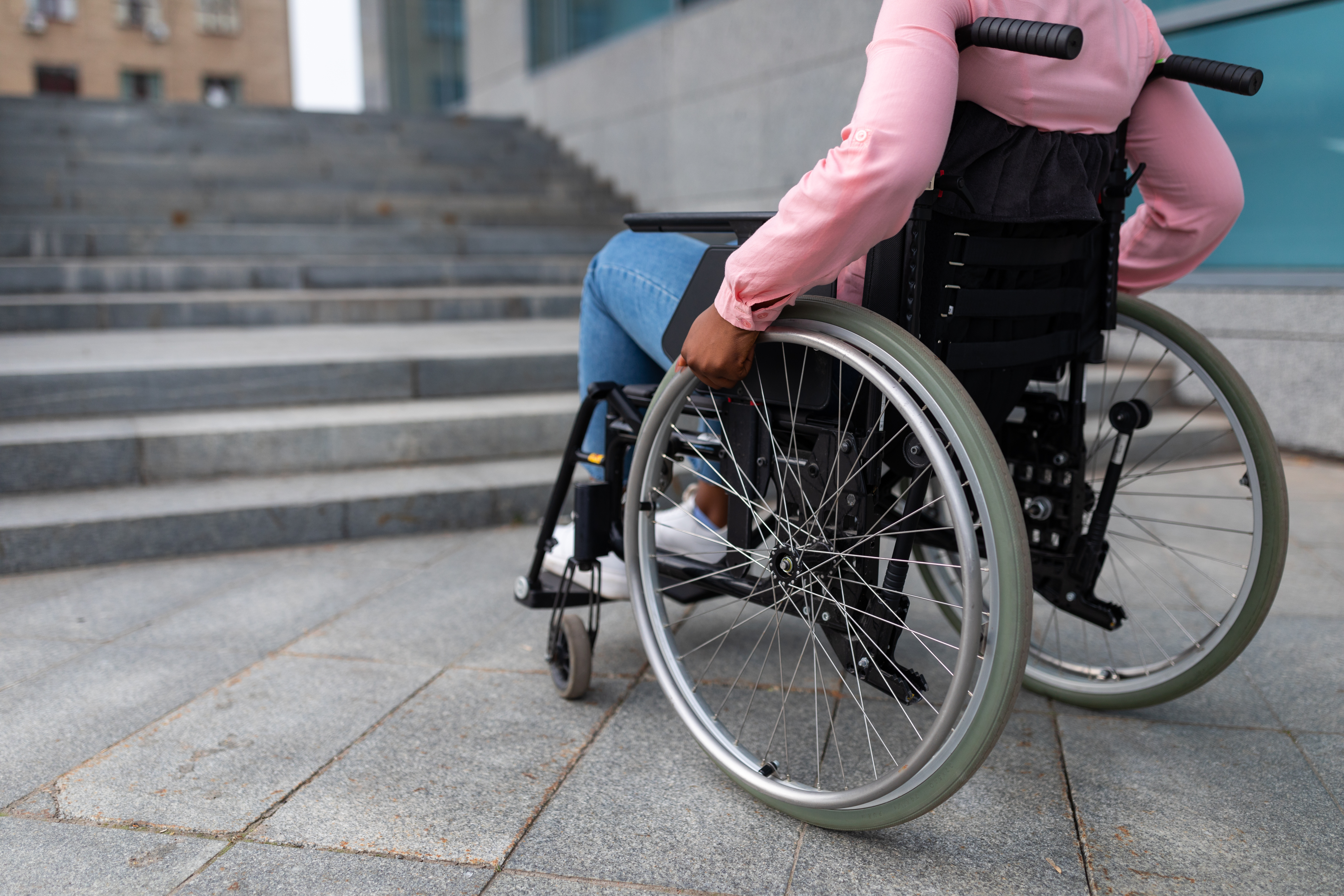 Black woman using a manual wheelchair at the base of a wide outdoor staircase beside a modern building,
