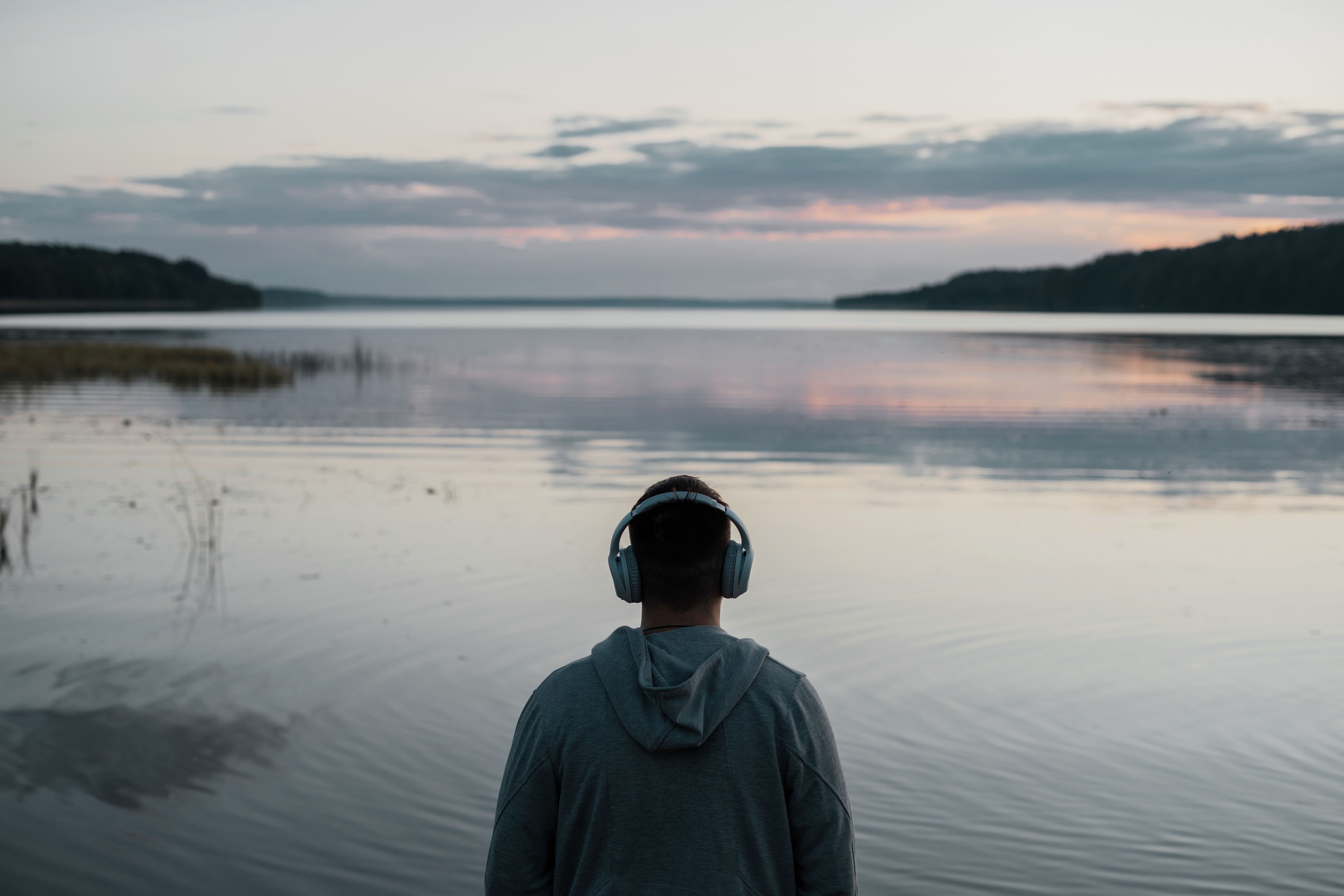 Man wearing over‑ear headphones stands facing a calm lake at dusk, with soft clouds reflected on the water