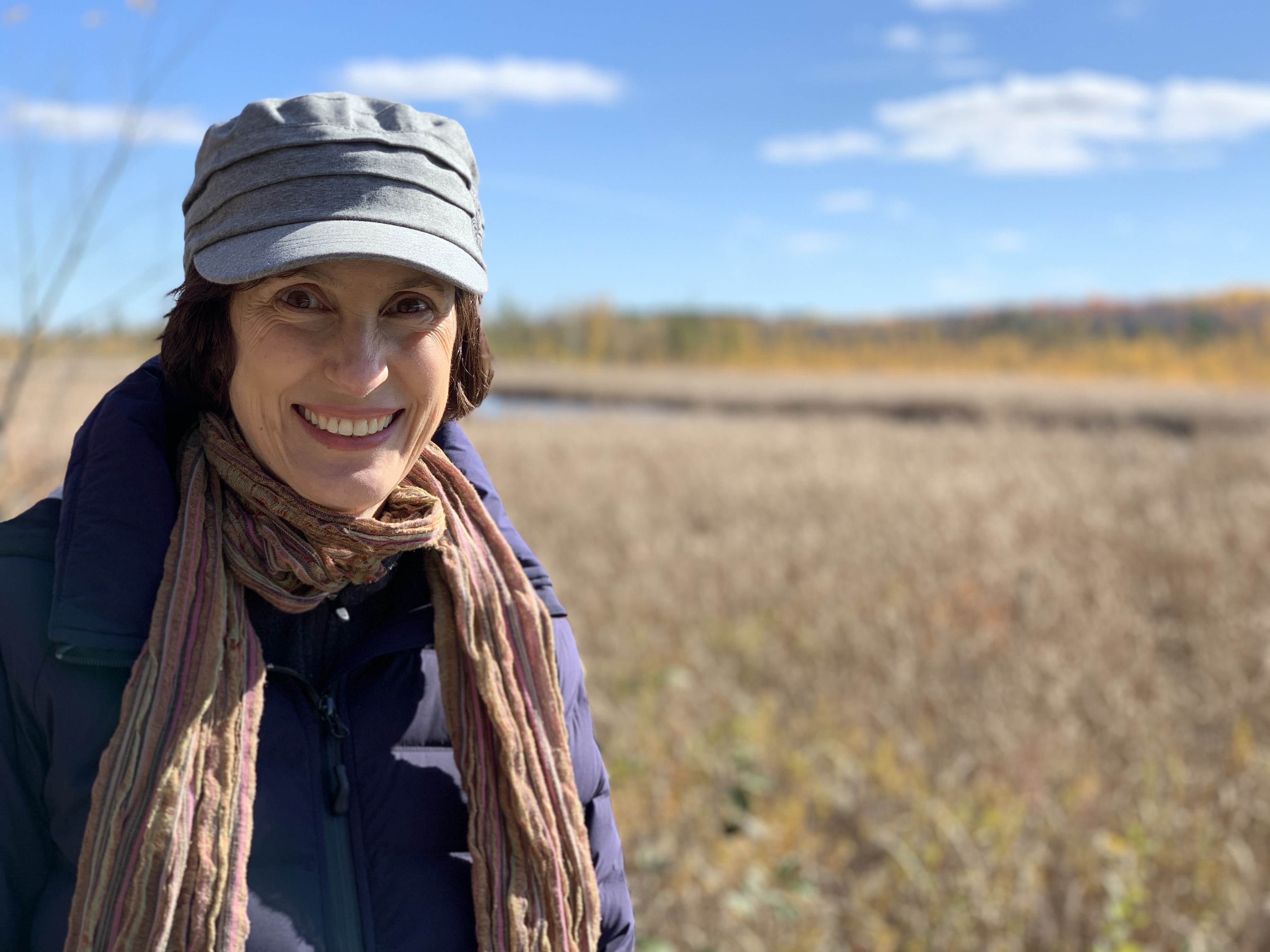 Smiling woman wearing a hat and scarf standing in a field on a sunny day