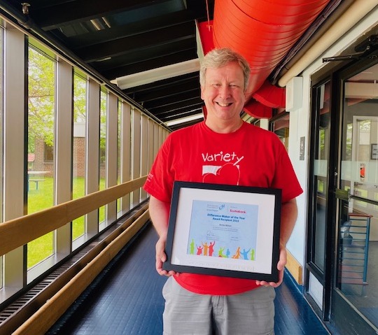 Man with short blonde hair wearing a red t-shirt. He is holding a framed Difference Maker of the Year award certificate. 