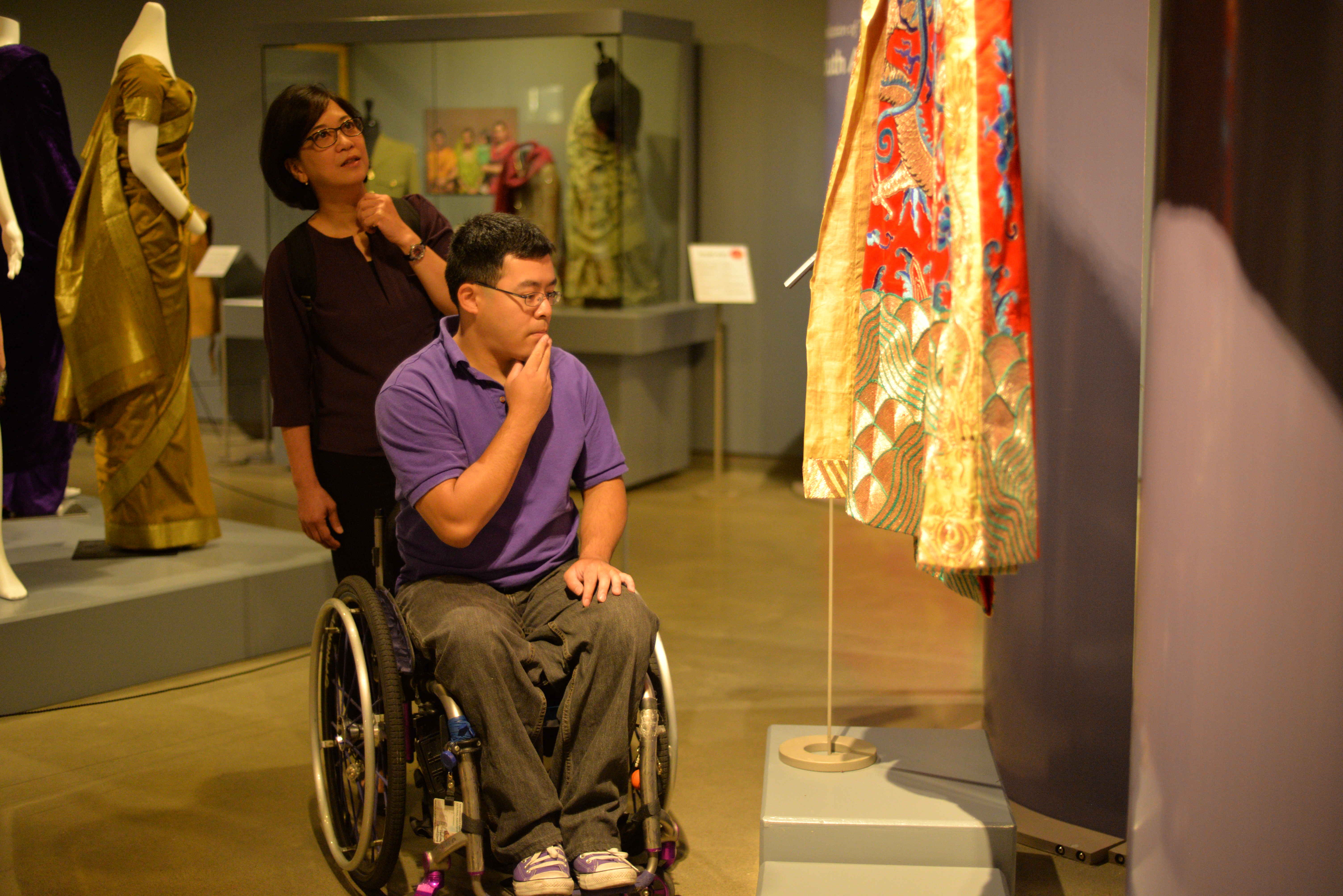 Man with black hair and glasses in a purple t-shirt using a wheelchair in a clothing exhibit. A woman wearing a brown shirt is standing behind him.