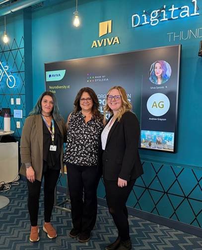 Three smiling women stand in front of a blue wall featuring the "Aviva" logo. A large TV screen to their side shows what appears to be a virtual meeting.