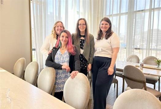 Four women are in a bright, elegant room with large windows and white furniture.