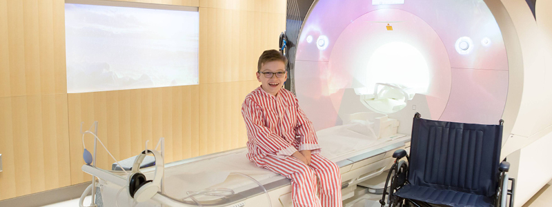 Child sitting on a bed of a medical imaging machine. There is a wheelchair beside him.