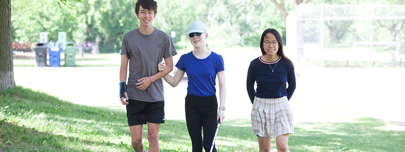 Three people walking. The person in the middle is using a white and red cane.