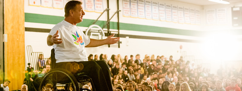 Rick hansen using his wheelchair on stage with his arms open wide looking at the crowd. He is wearing a white t-shirt with a colourful logo.