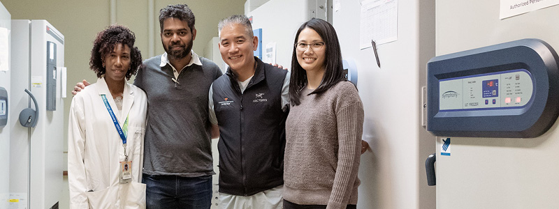 A man and woman with dark skin are standing next to an Asian man and woman in front of a lab. The black woman is wearing a lab coat. 