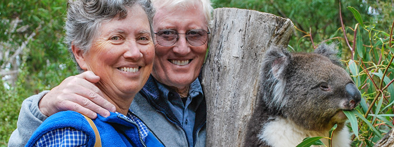 An elderly white man and woman smiling next to a koala. 