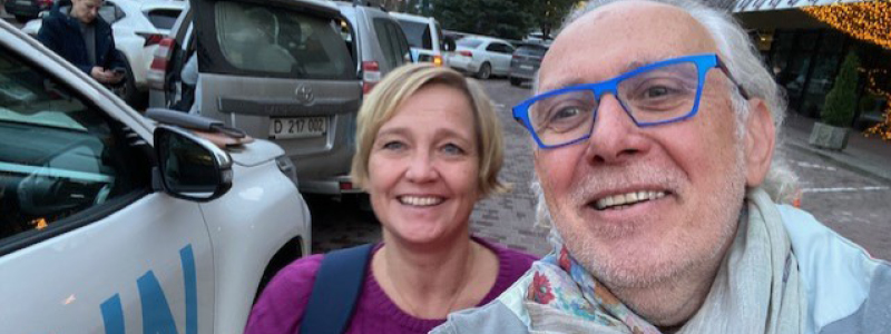 Two people taking a photo together, selfie-style. One person has bright blue glasses and short grey hair. The other is wearing a purple shirt and has ear-length blonde hair.