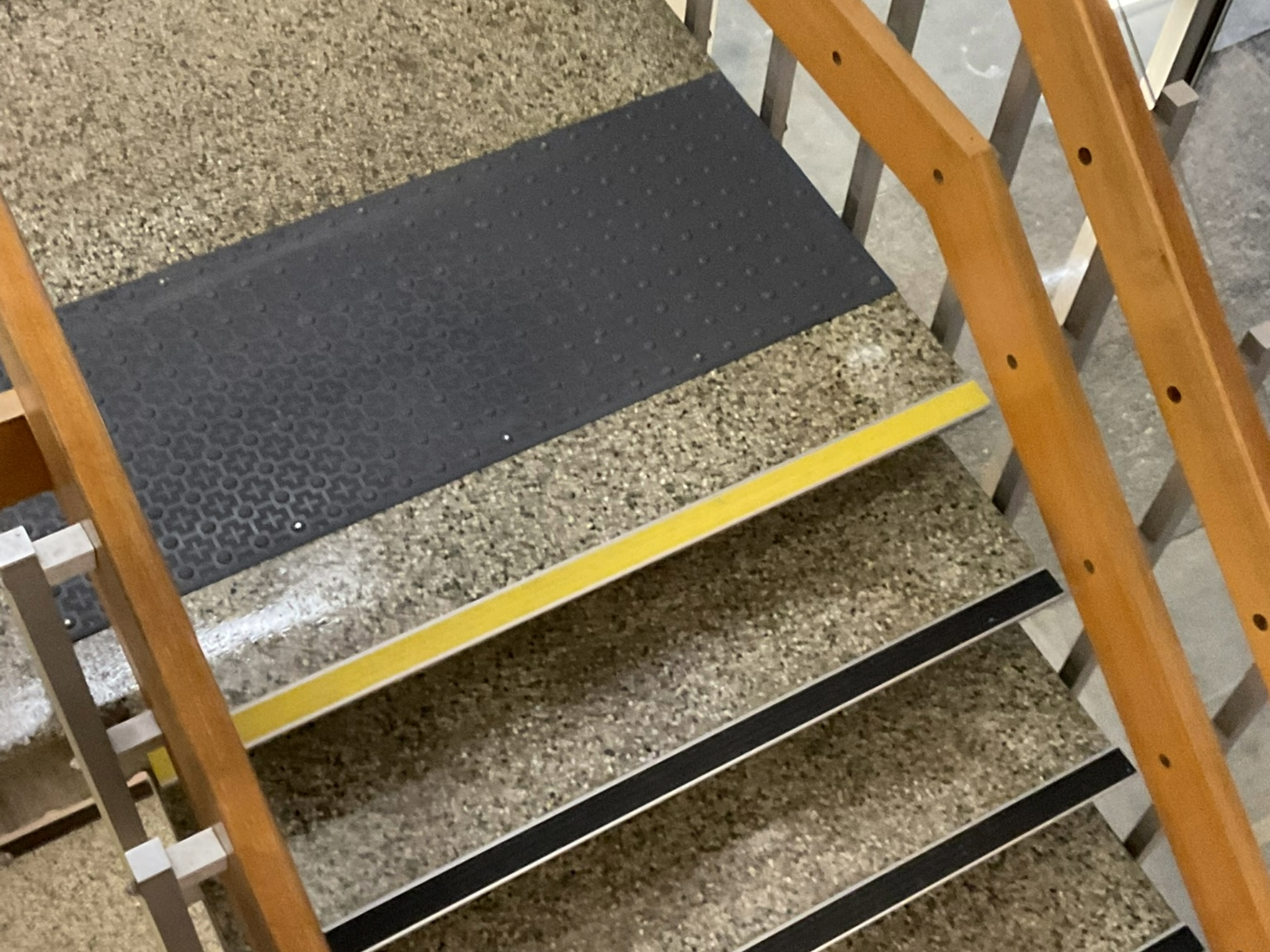 An indoor staircase with textured steps. The top landing has a black tactile strip with raised bumps, and the stair edges are marked with yellow and black anti-slip strips.