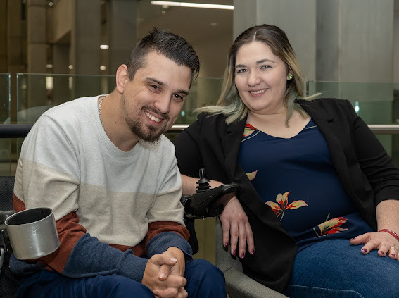 Brad and his wife who are facing the camera and smiling. Brad is using a wheelchair.