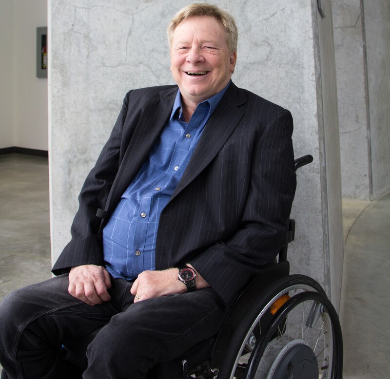 Brad McCannell who has short, light-coloured hair and is wearing a blue button up shirt and a dark sports coat. He is in front of a grey pillar and is using a wheelchair.