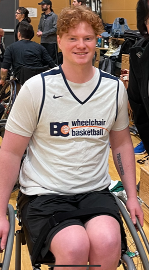 Cameron smiling at the camera. He has red hair and is wearing a BC wheelchair basketball jersey. Cameron uses a wheelchair.