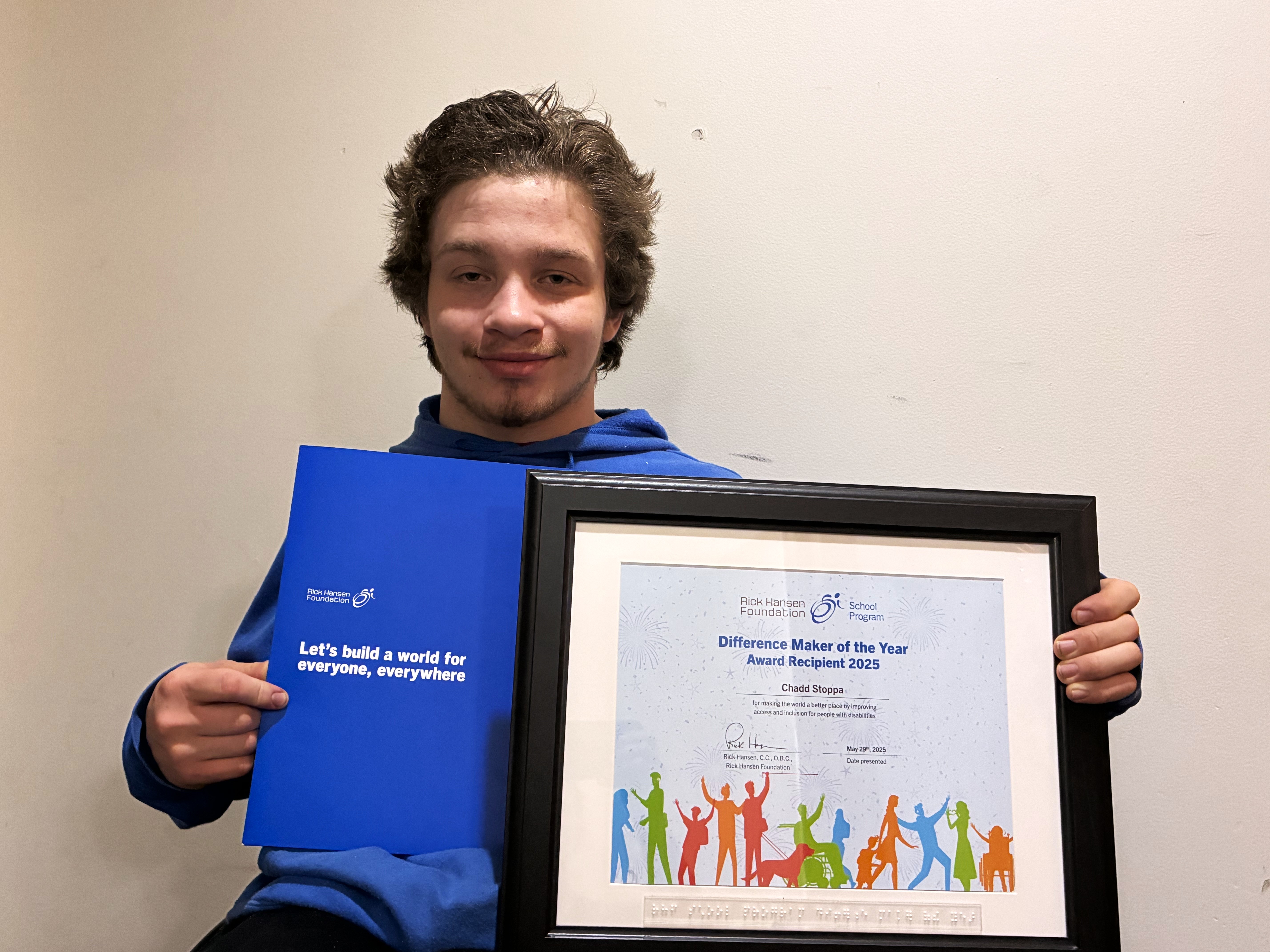 Jeune homme blanc souriant, tenant un certificat « Lauréat du prix Artisan du changement 2025 » et une pochette bleue avec l’inscription « Construisons un monde pour tous, partout »