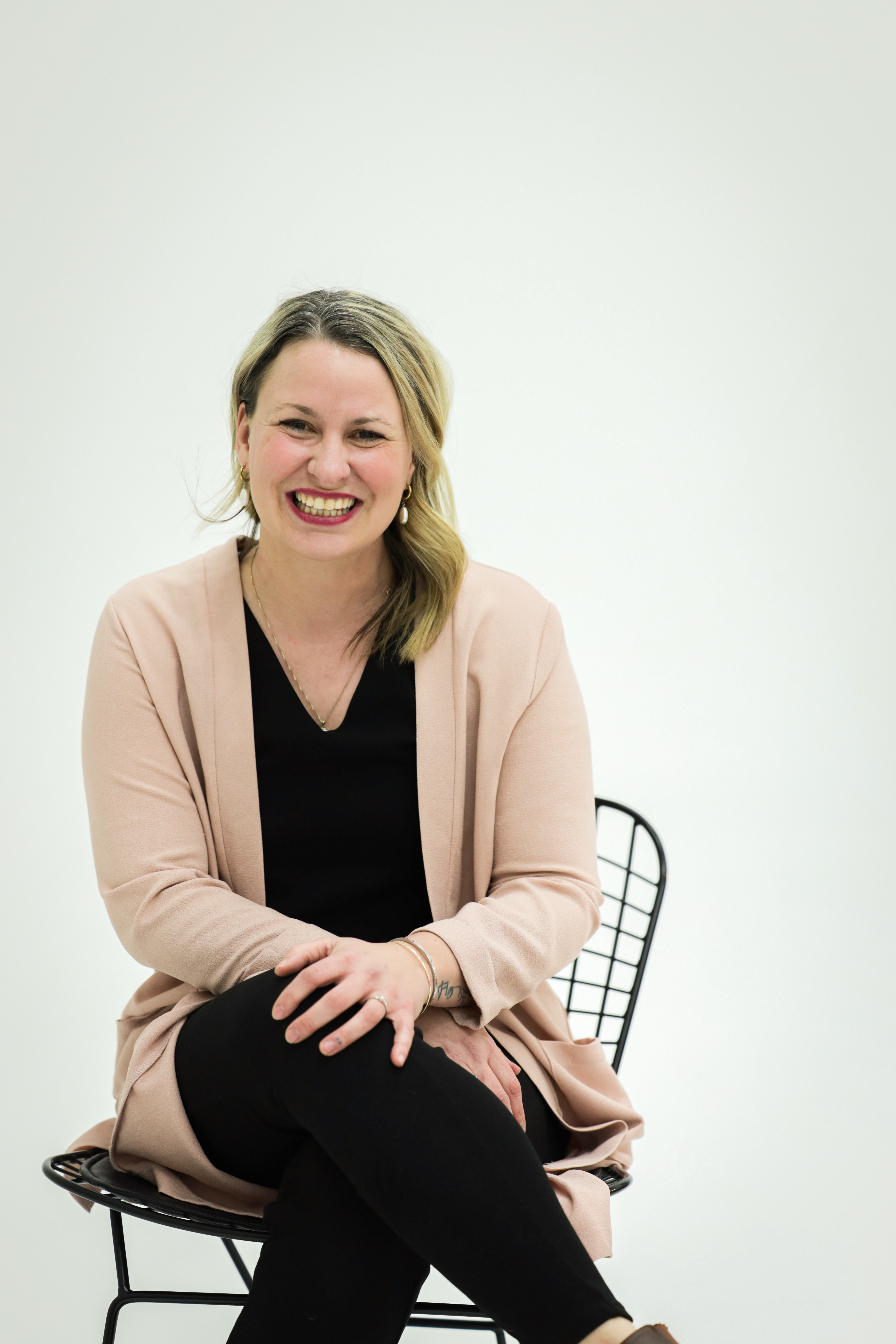 A woman seated on a chair against a plain light background. She has one leg crossed over the other and hands resting on the knee. 