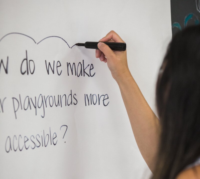 Person with long dark hair writing on a whiteboard in black marker. The writing reads "how do we make playgrounds more accessible?"